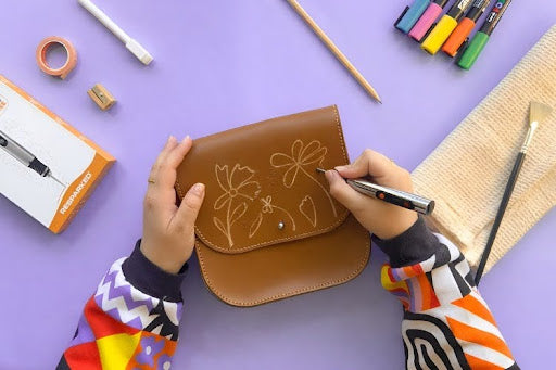 Person engraving floral designs onto a leather bag using a handheld engraving pen.
